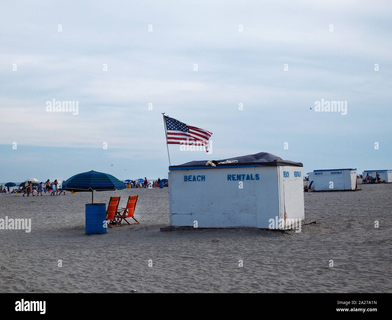 Small wooden shacks spraypainted with stenciiled blue letters reading 'BEACH RENTALS' line an American beach on a busy summer day at the seaside. Stock Photo