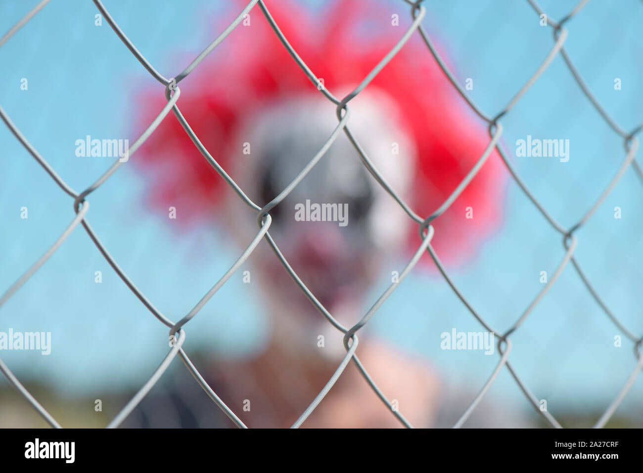 closeup of a chain-link fence in focus, and a blurred scary clown in the background Stock Photo