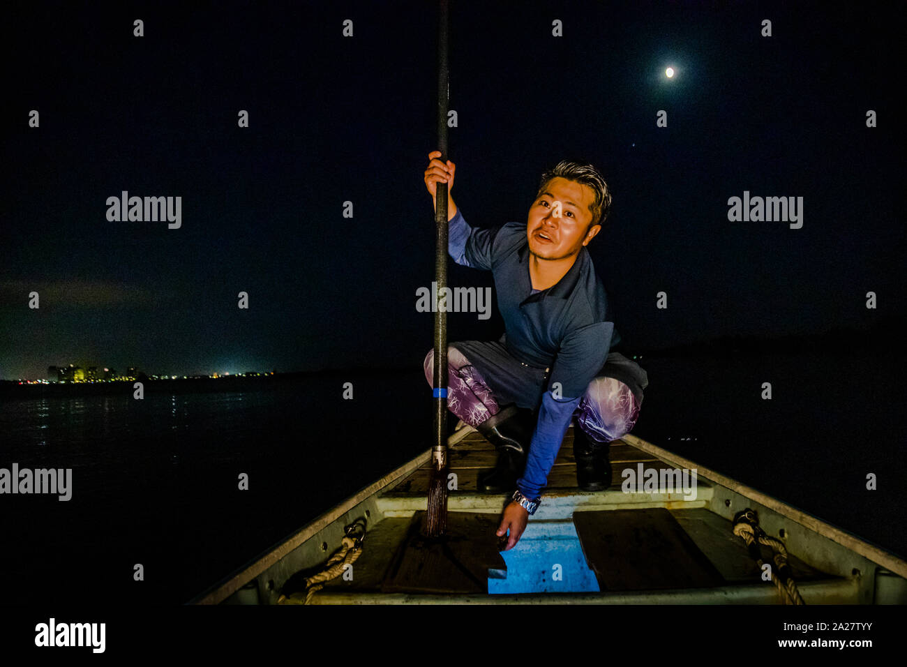 Fisherman Ikuya Yamamoto crouches at the bow of his boat on Lake Hamana near Hamamatsu, Japan, pointing towards the water, the full moon hanging low over the dark shoreline. Stock Photo
