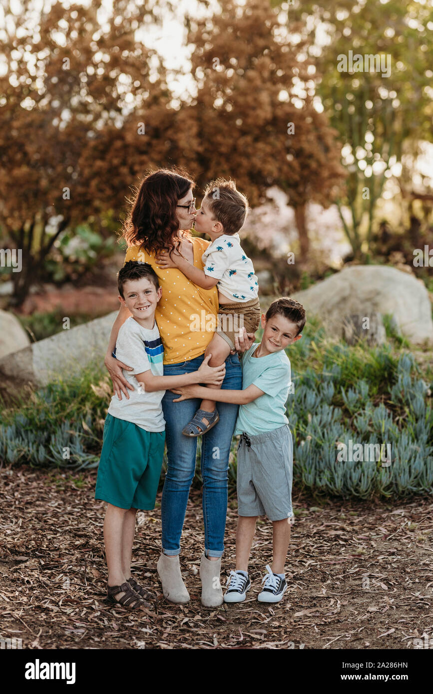 Portrait of mother kissing son with boys hugging her in sunny garden Stock Photo