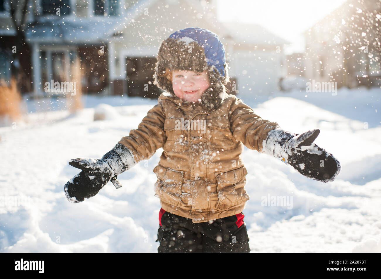 Toddler boy playing and smiling while throwing snow in front of house Stock Photo