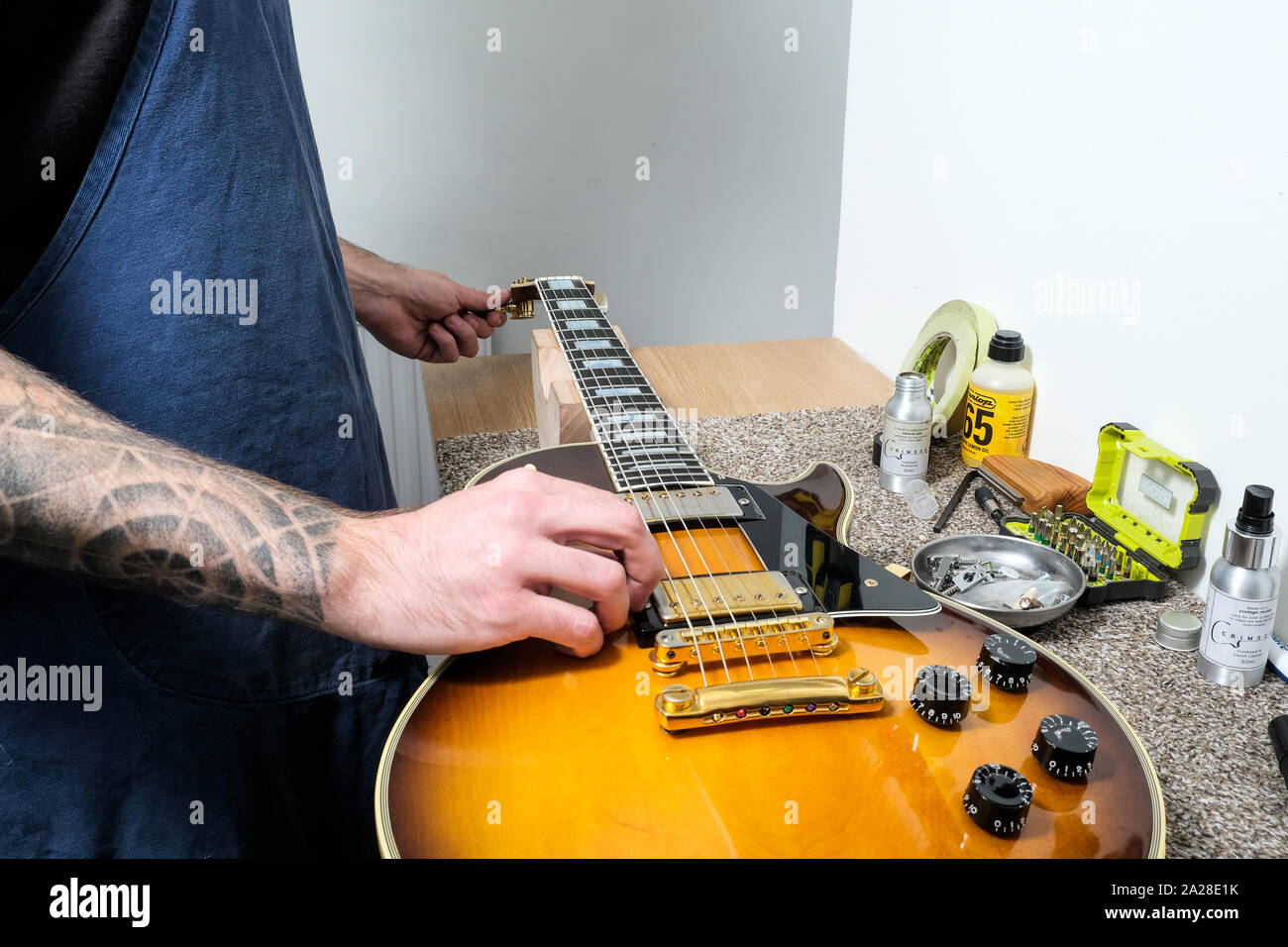 A guitar technician or tech tuning a re strung Gibson electric guitar in his workshop. He's turning the tuning post whilst plucking the string Stock Photo