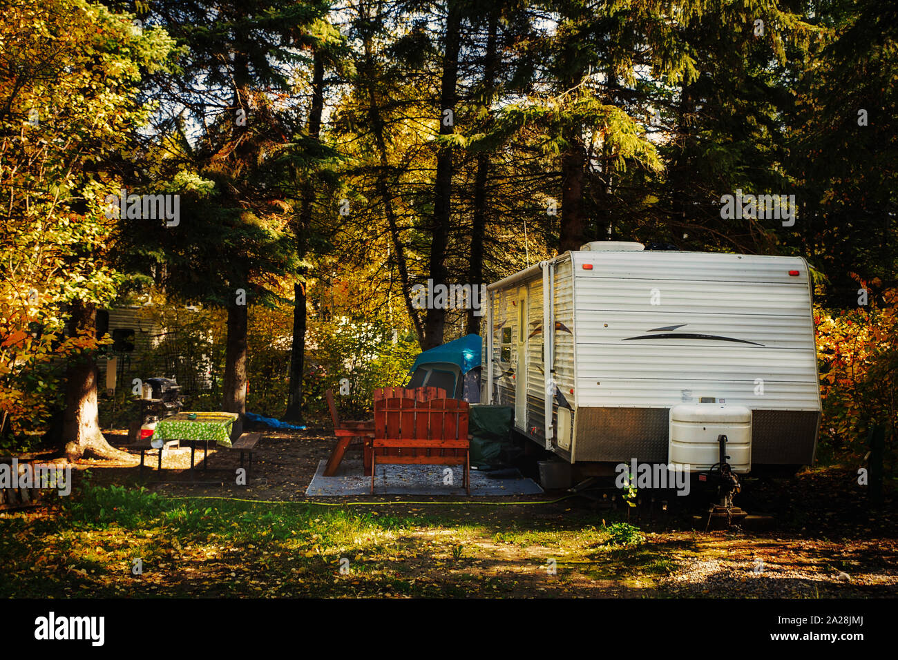 A deserted camper trailer and tent with wooden chairs on outdoor carpet and cloth covered table surrounded by tall pine trees parked in a deserted cam Stock Photo