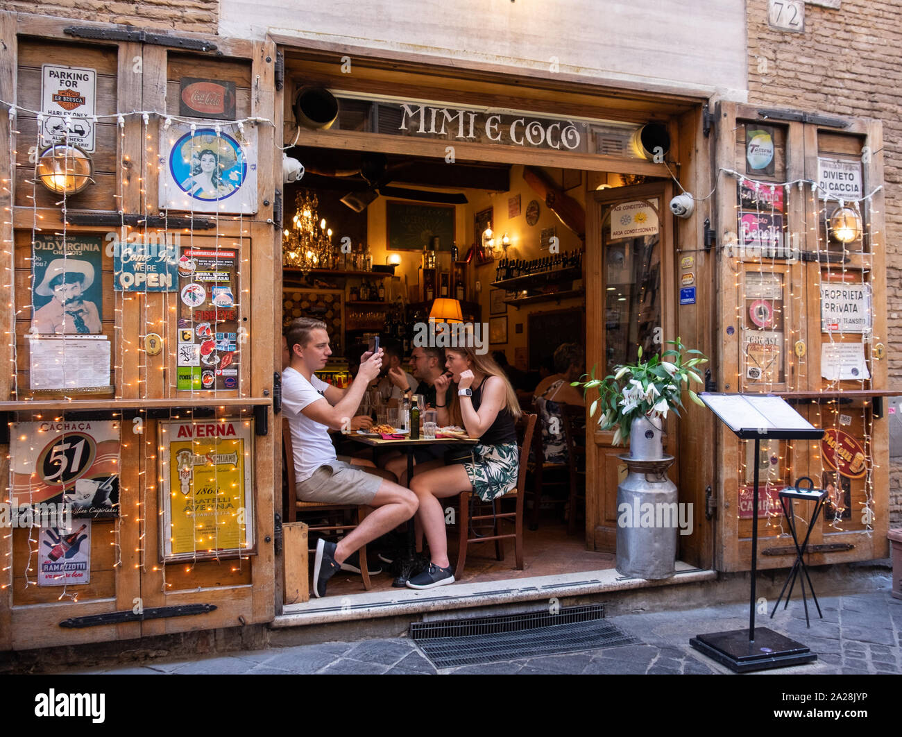 Young couple using phone sat over lunch in traditional street side cafe in Rome, surrounded by coloured posters and lights. Stock Photo