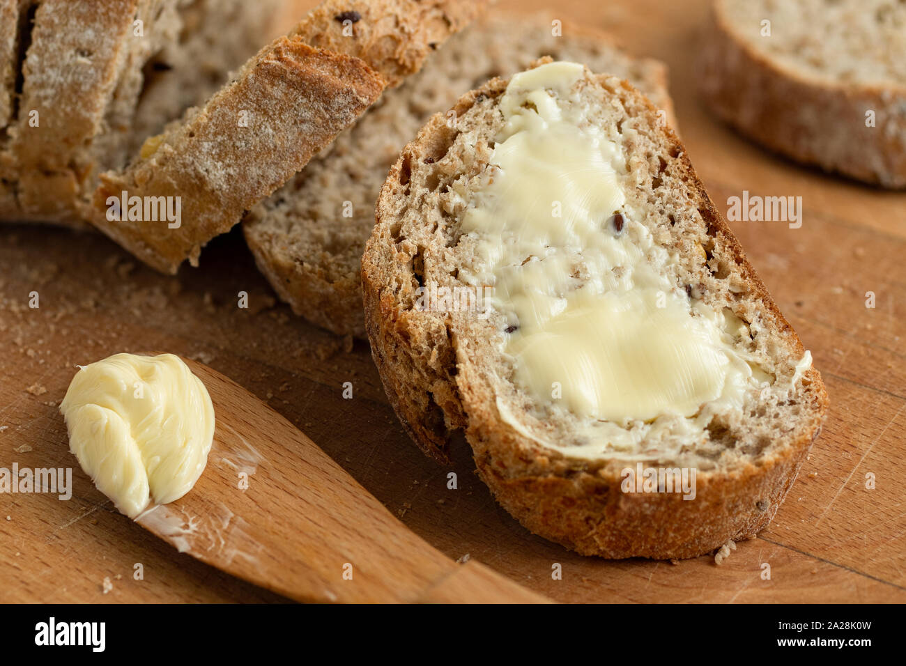 Closeup of buttered slice of whole wheat rustic bread next to cut bread and wood knife. Light wood board. Stock Photo