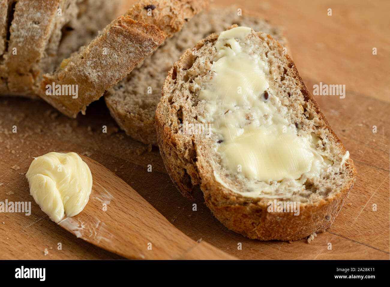 Closeup of buttered slice of whole wheat rustic bread next to cut bread and wood knife. Light wood board. Stock Photo