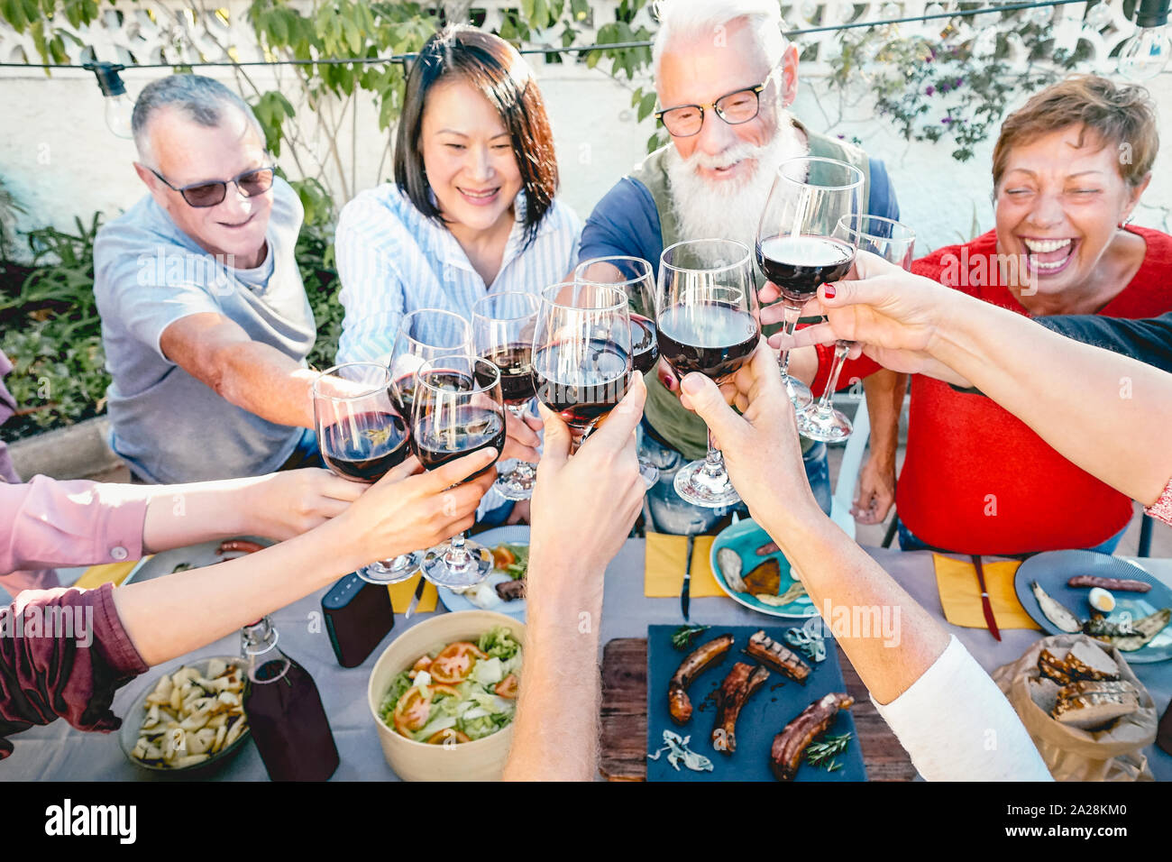 Happy senior friends toasting with red wine glasses at dinner time outdoor - Mature people having fun dining together outside Stock Photo