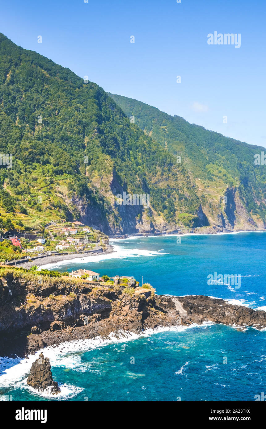Beautiful cliffs on the coast of Atlantic ocean by village Seixal, Madeira Island, Portugal. Steep green rocks, small city and blue sea water captured on vertical photography. Tourist destination. Stock Photo