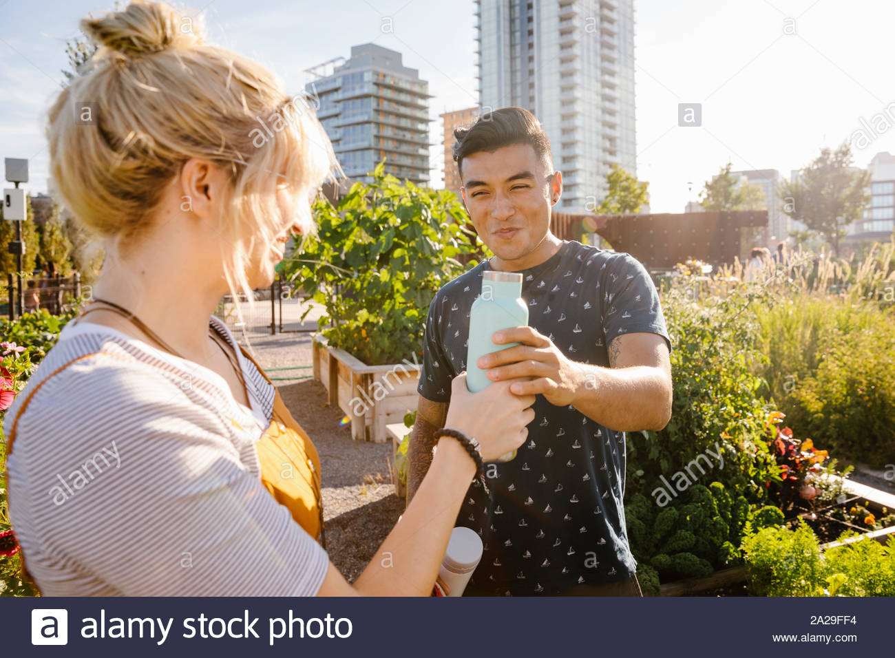 Happy young couple sharing water bottle in sunny, urban community garden Stock Photo