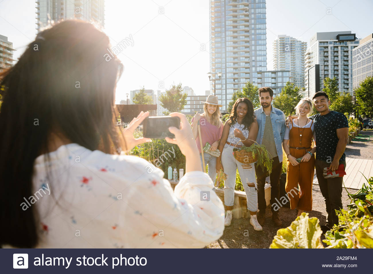 Young woman with camera phone photographing friends in sunny, urban community garden Stock Photo