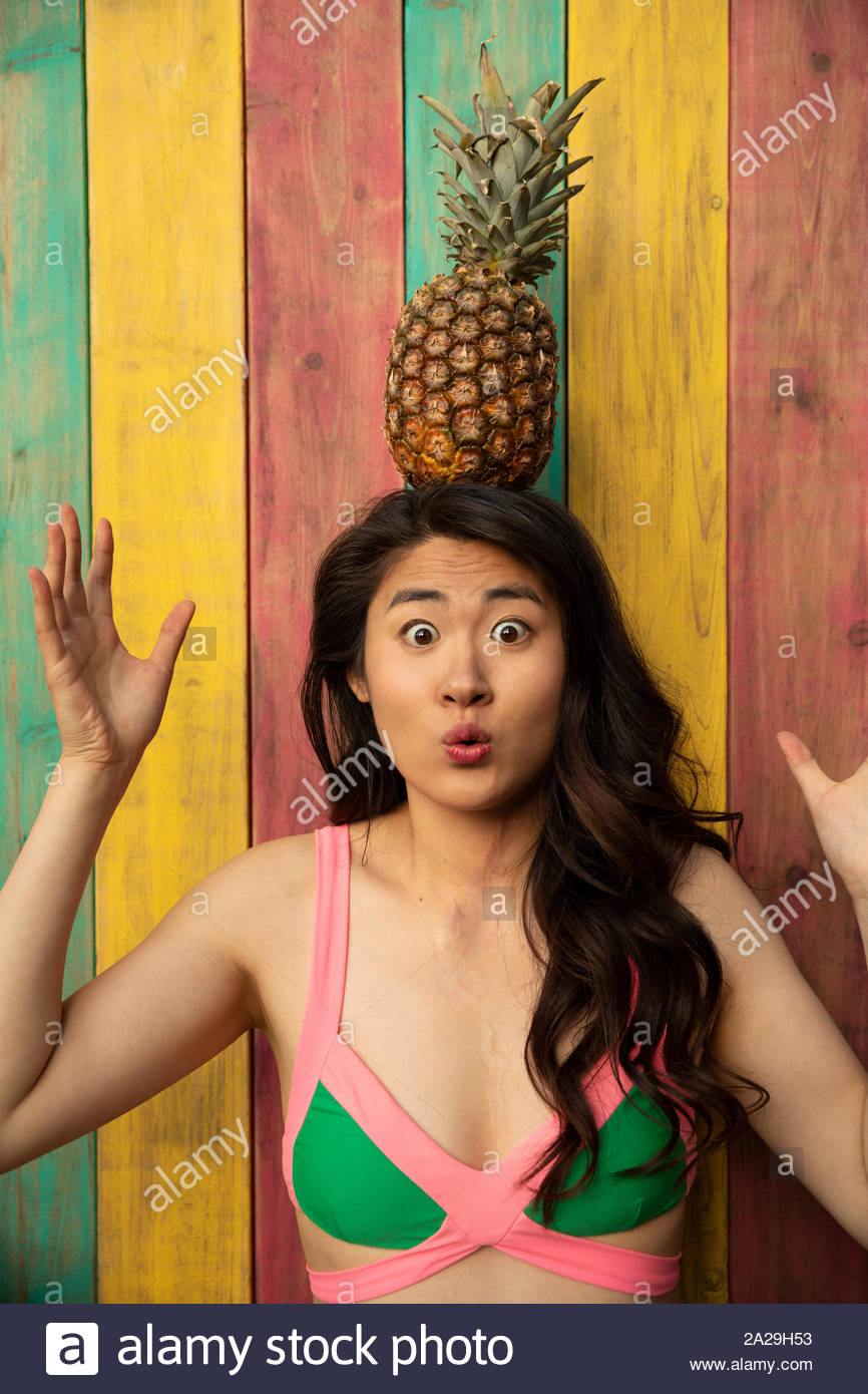 Portrait playful, wide-eyed young woman in bikini top balancing pineapple on head on summer patio Stock Photo