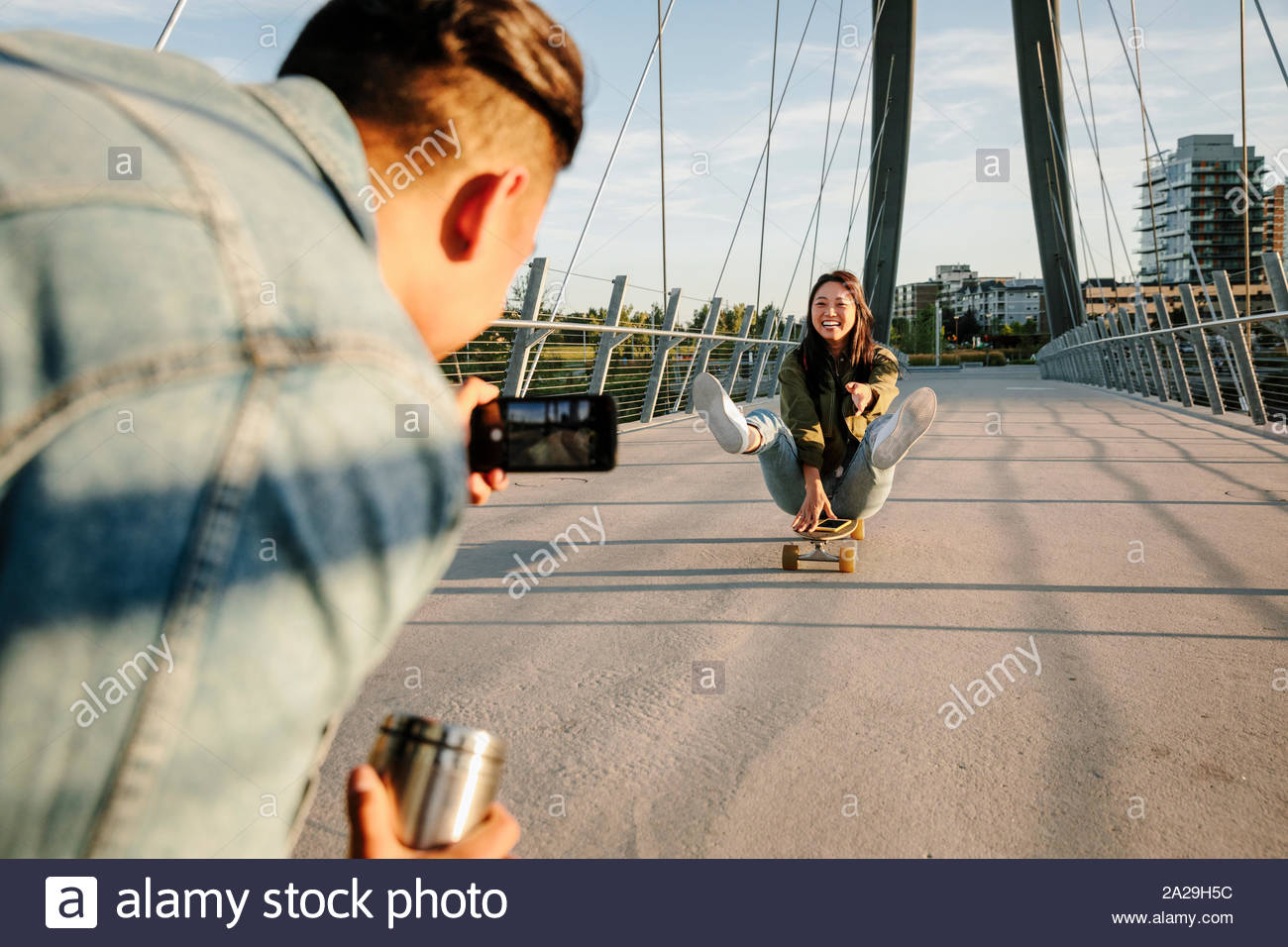 Young man with camera phone photographing playful girlfriend on skateboard Stock Photo