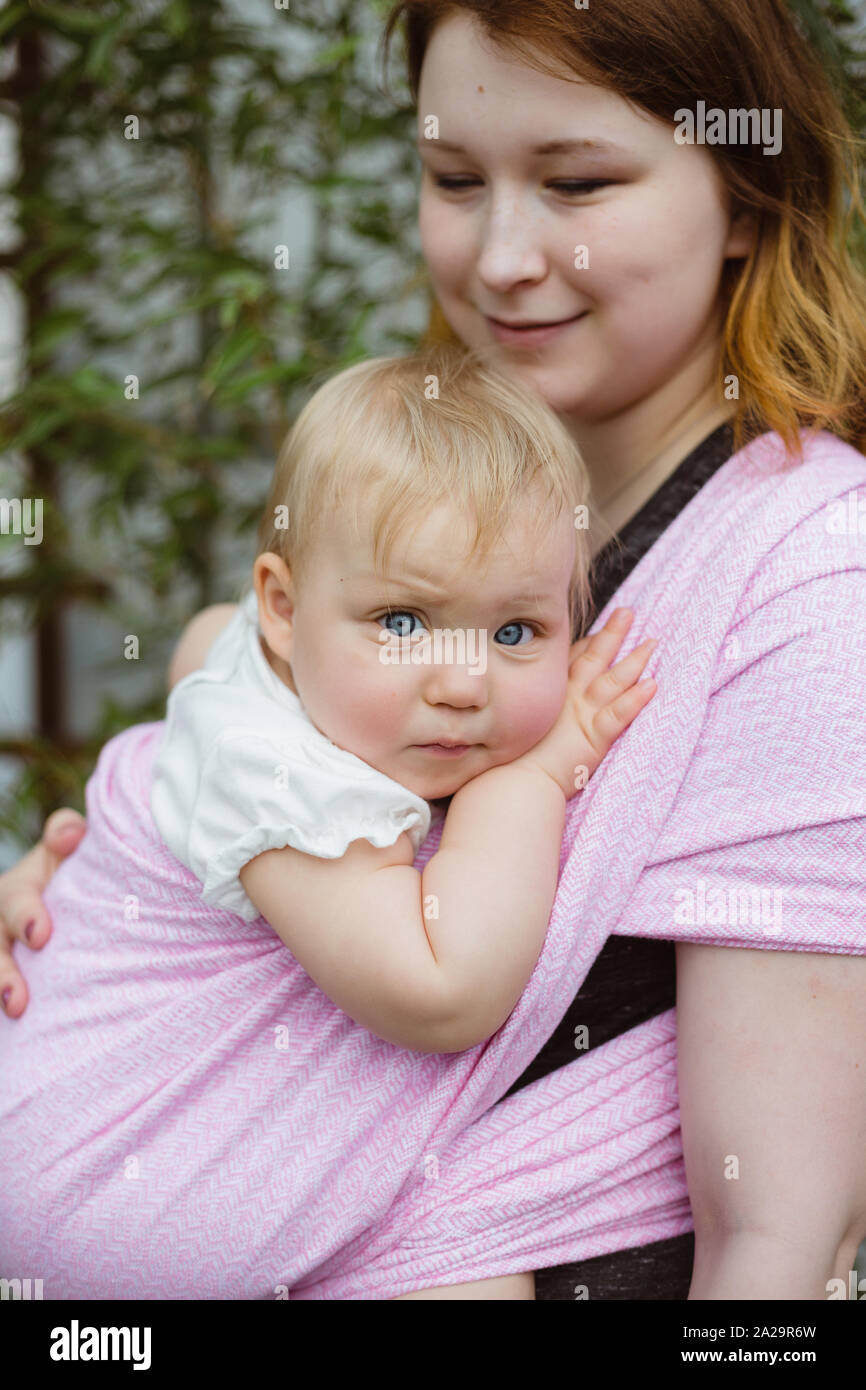 Young redhead babywearing mother carry her sleepy one year old baby girl in a pink wrap sling Stock Photo