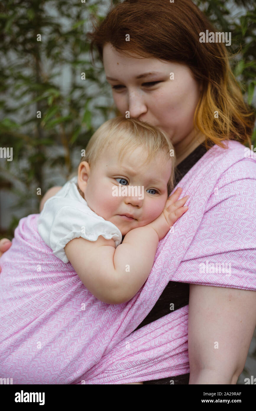 Young redhead babywearing mother carry her sleepy one year old baby girl in a pink wrap sling