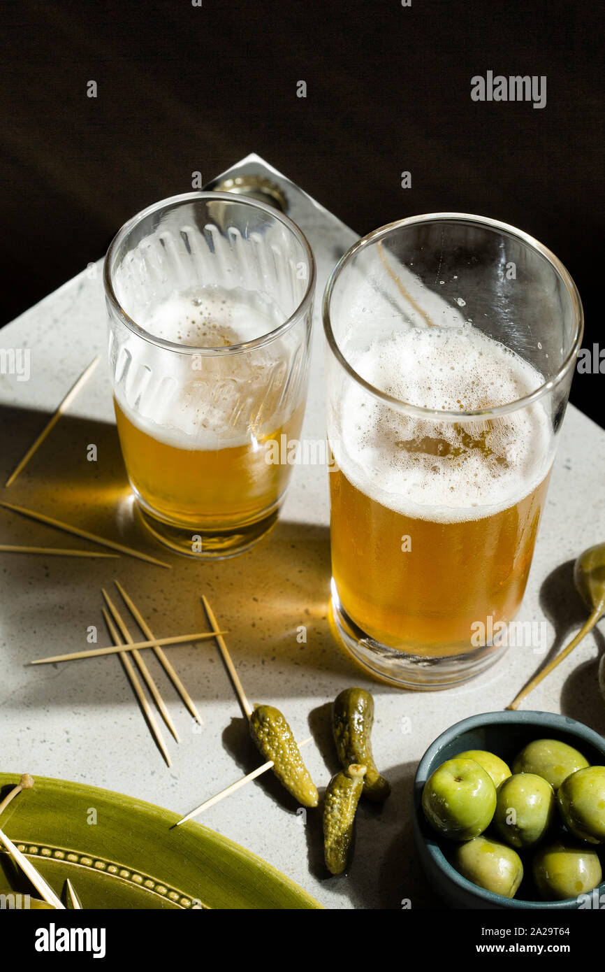 Beer and snacks. A selection of pickled gherkins, caper berries and green olives accompany two glasses of beer on a concrete background. Stock Photo