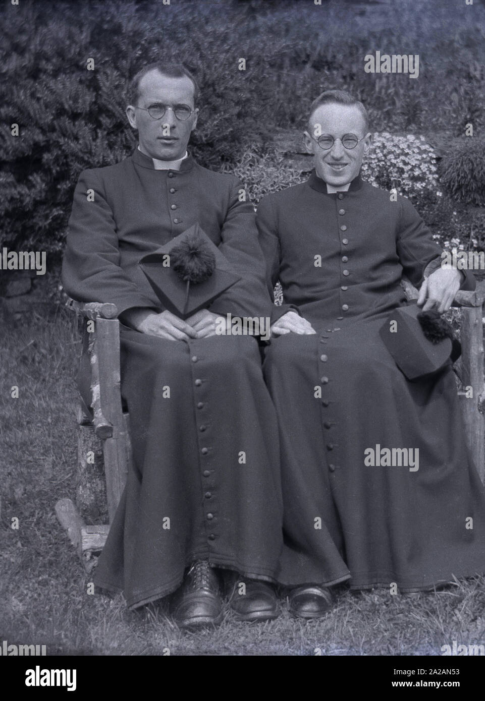 circa 1920s, historical, two Roman catholic priests sitting outside in their long robes on a small bench, holding their ecclesiastical hats, a biretta, a stiff square cape wth four ridges across the top, surmounted by a tuft, England, UK. The long button ankle-length gowns are known as cassocks and are a traditional christian clerical clothing coat. Stock Photo