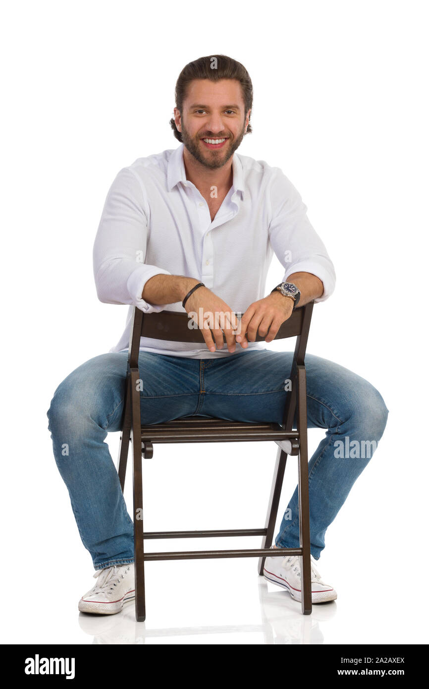 Handsome man is sitting on a chair, looking at camera and smiling. Front view. Full length