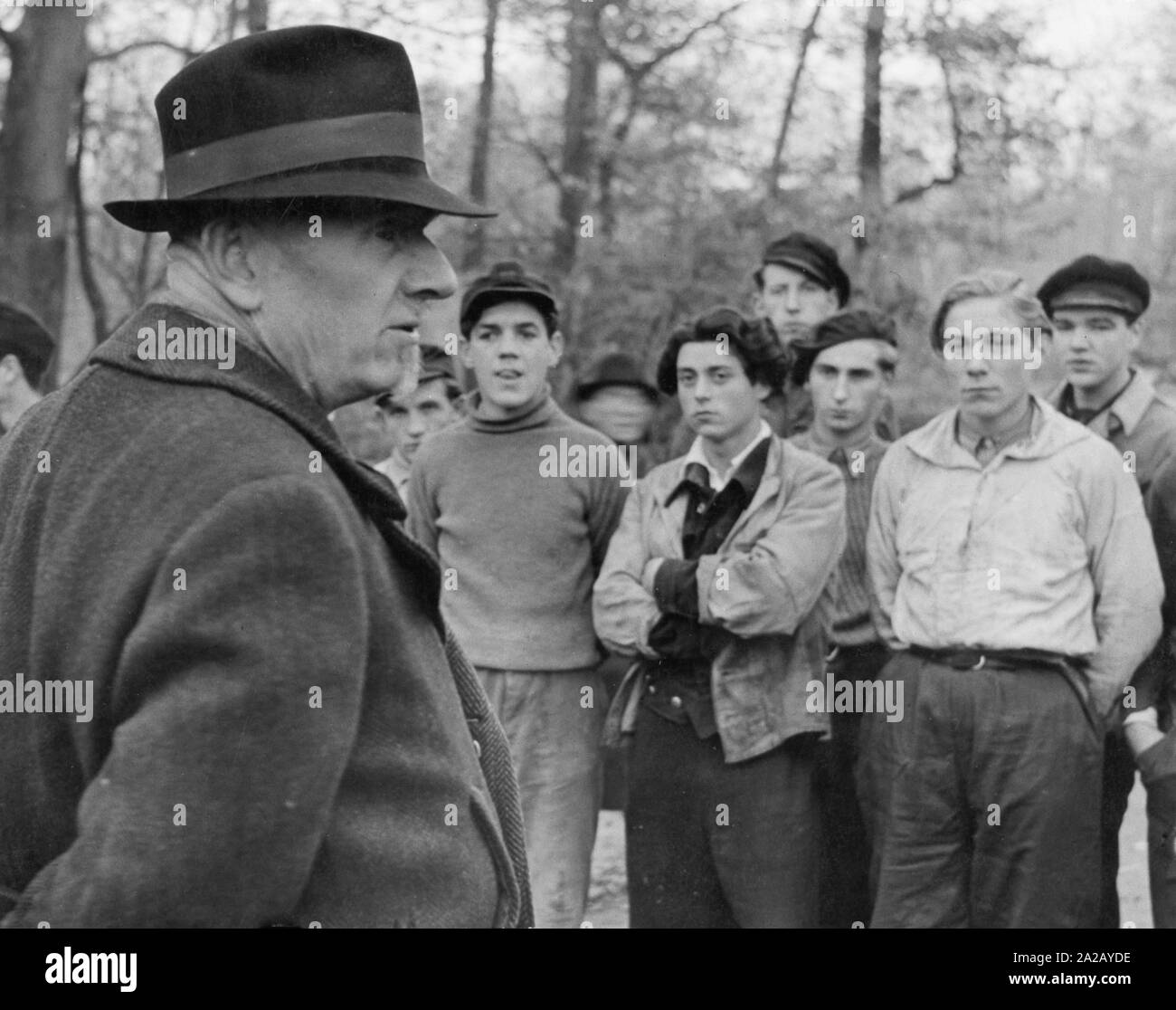 An elderly man stands in front of a group of young men. Two generations face each other suspiciously. Undated picture. Stock Photo