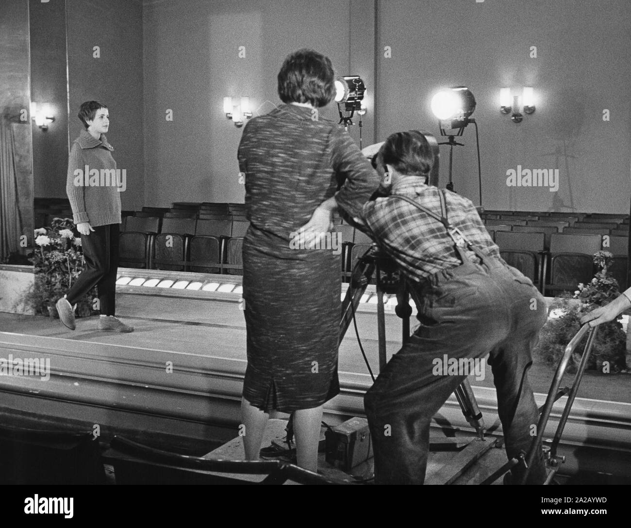 A model walks on the catwalk in everyday clothes, while two employees check the lights and camera. Stock Photo