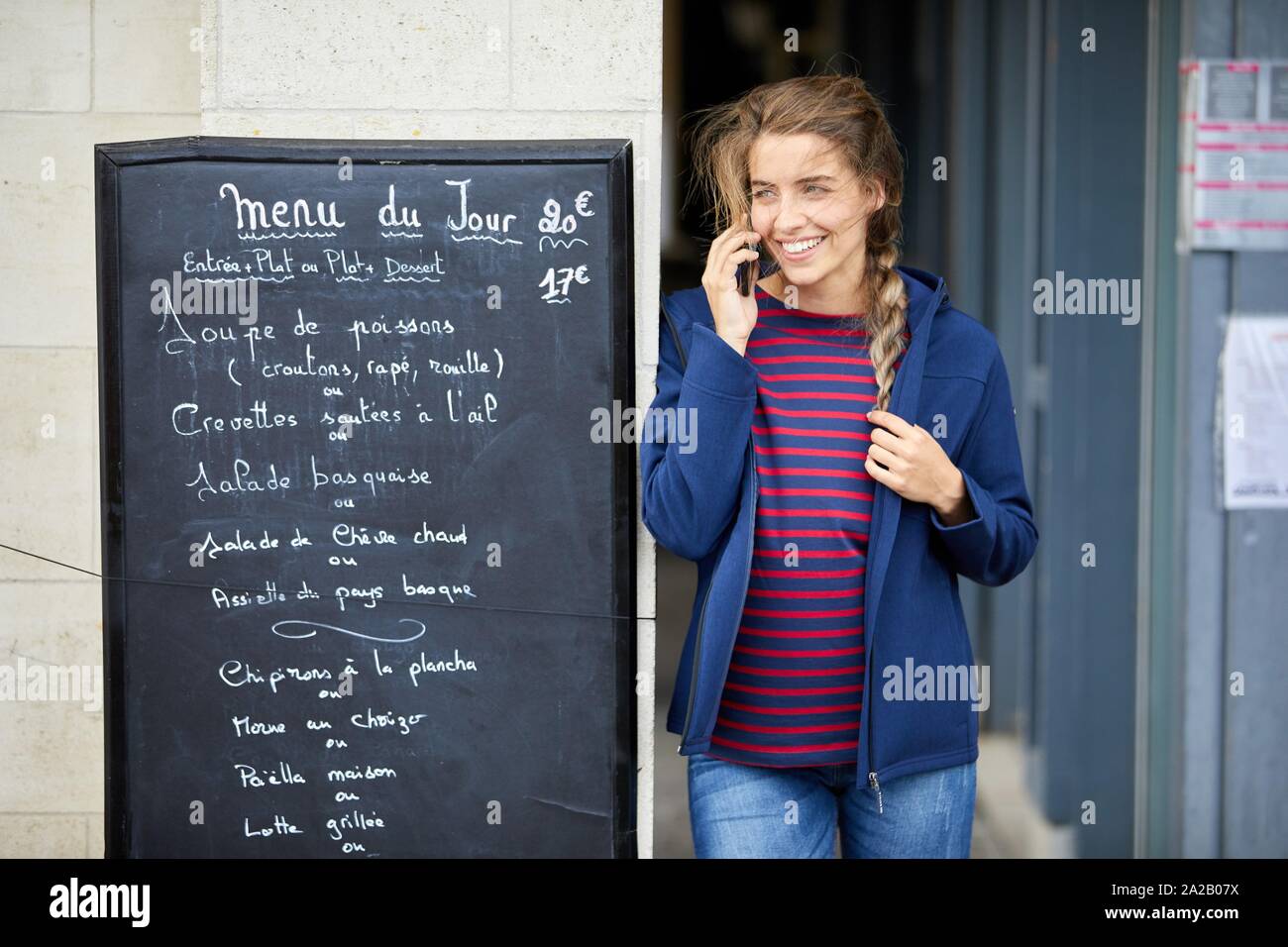 Woman beside restaurant menu, talking on phone. Sokoburu, Hendaye, Aquitaine, Basque Country, France Stock Photo