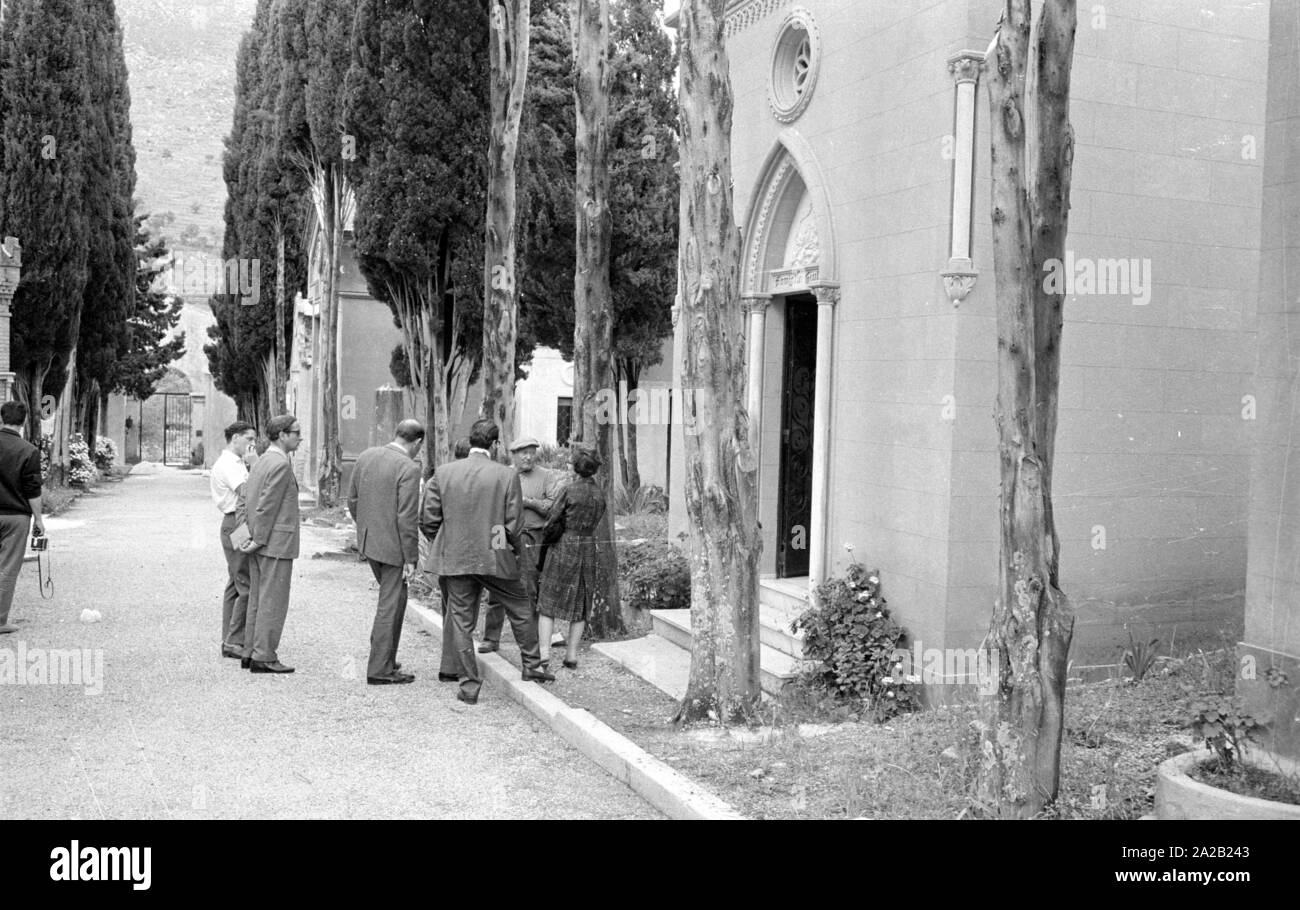 Photo of some visitors and reporters in the Ostfriedhof of the town of Montelepre in the province of Palermo (region of Sicily). A local man is talking to the visitors in front of the tomb. Former folk hero Salvatore Giuliano (also born in Montelepre) is buried in the cemetery. He was long regarded as a kind of 'Sicilian Robin Hood', but he also had connections with the mafia. Stock Photo