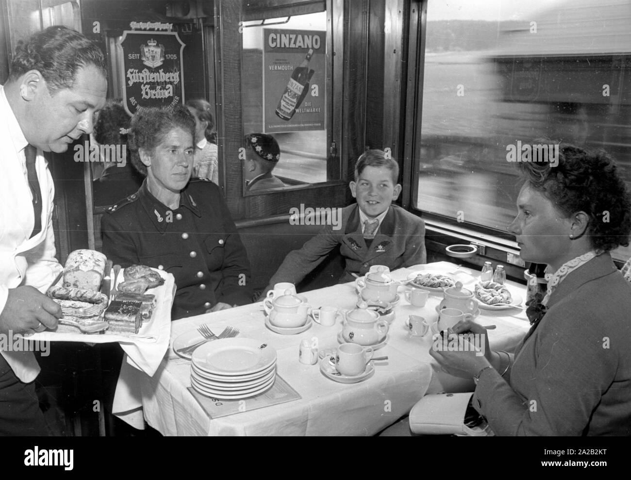 Photo of the interzonal train on the route Leipzig-Gutenfuerst-Hof-Munich. This train was known for its 'all-German' dining car, which was popular due to the 1: 1 exchange rate, especially among East German travelers. Photo of train passengers drinking coffee and eating cake in the dining car. The East German railway employee (left) with her two children sits at the table together with a woman from Munich and a student from the village. A waiter brings another tray of cake. Stock Photo