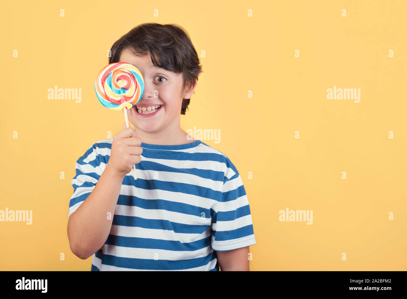 funny child covering eye with lollipop on yellow background Stock Photo