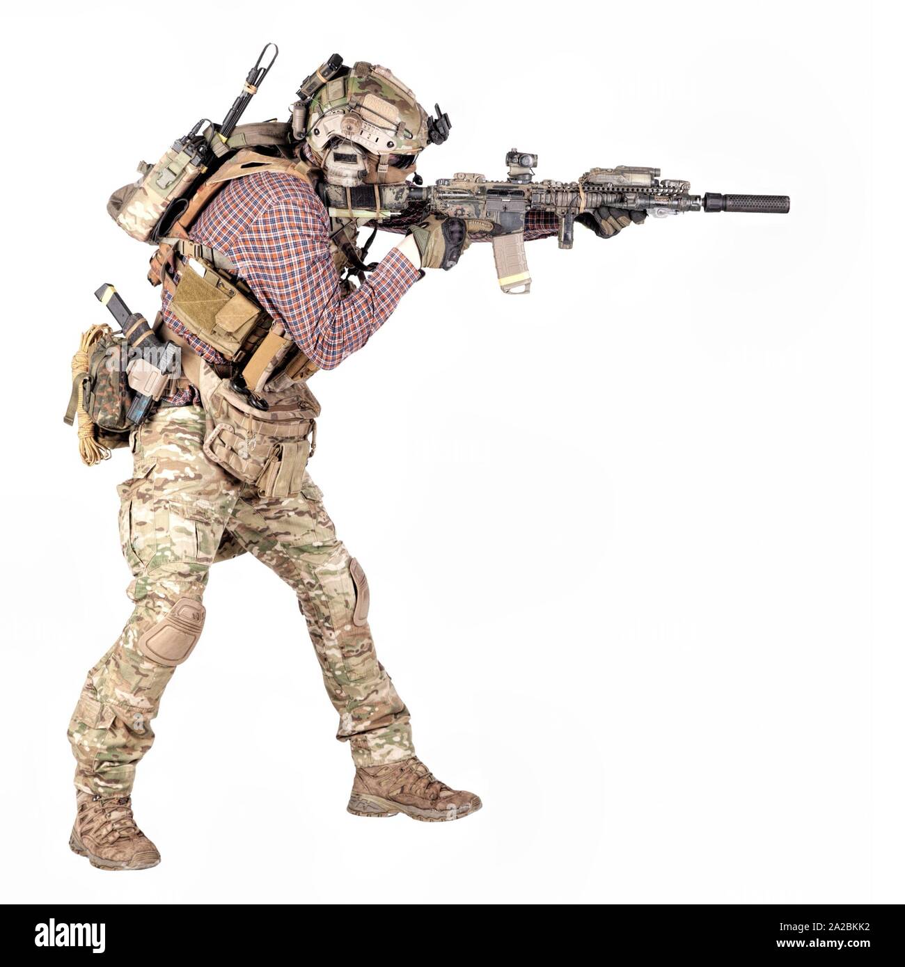 Full length portrait of airsoft player in checkered shirt, wearing camouflage uniform, helmet with tactical radio headset, body armour, aiming with Stock Photo