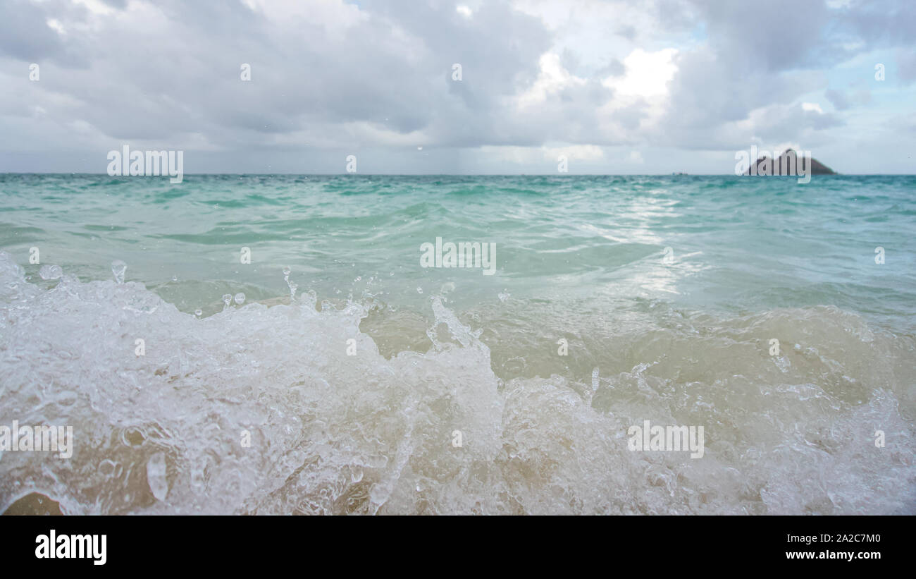 Close up of wave crashing onto beach with small islands out of focus on the horizon on an overcast sky. Stock Photo
