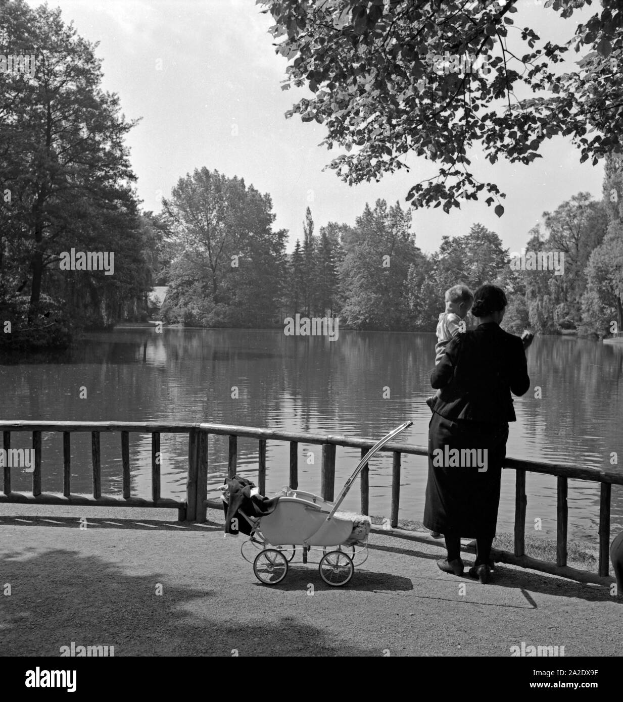 Eine Frau hat ihr Kind aus dem Kinderwagen auf den Arm genommen und steht mit ihm am Ufer eines Sees in einem Park in Plauen, Deutschland 1930er Jahre. A woman with her child on her arm at the shore of a lake in a park at Plauen, Germany 1930s. Stock Photo