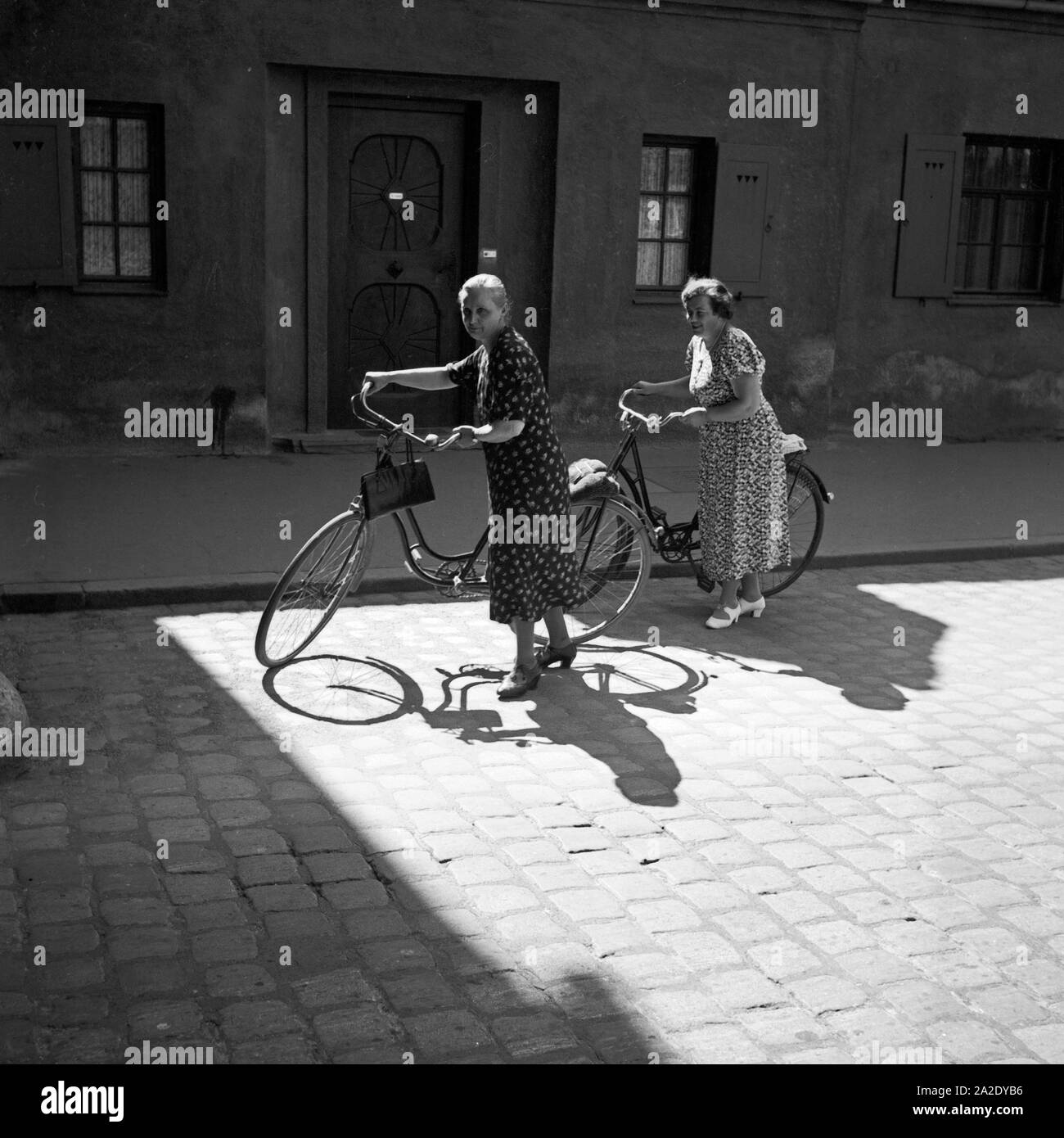Zwei Damen schieben ihre Fahrräder über das Kopfsteinpflaster von Augsburg, Deutschland 1930er Jahre. Two ladies pushing their bicycles through the old city of Augsburg, Germany 1930s. Stock Photo