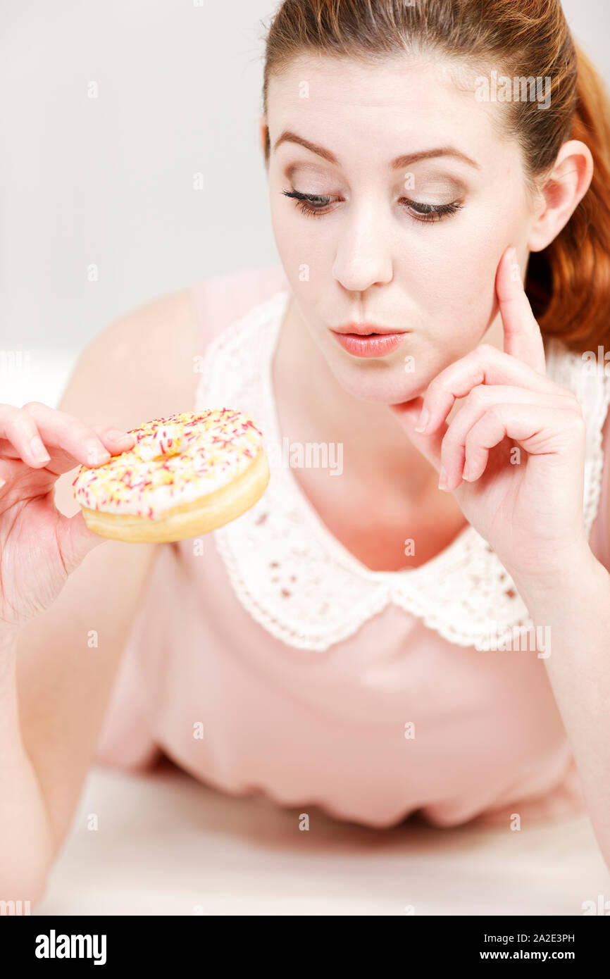 Young woman deciding whether to eat an unhealthy doughnut expressing guilt. Stock Photo