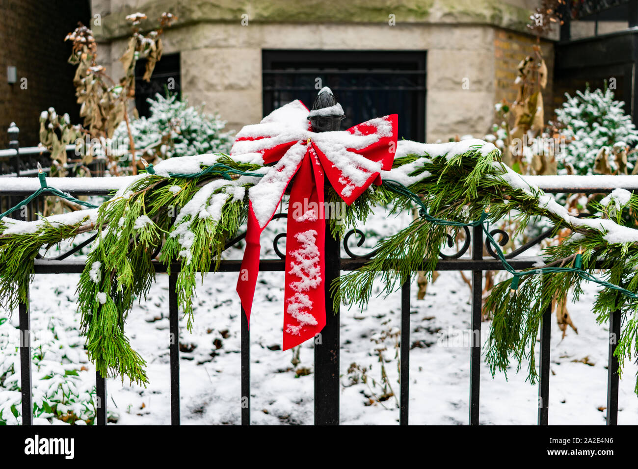 Red Holiday Bow on a Home Garden Fence wrapped with a Pine Garland and Lights during Winter with Snow Stock Photo