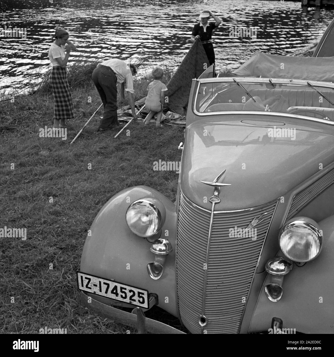 Eine Familie baut ein Zelt auf neben einem Ford V8 Cabrio, am Ufer eines Sees, Deutschland 1930er Jahre. A family bringing up a tent beside their Ford V8 convertible at the shore of a lake, Germany 1930s. Stock Photo