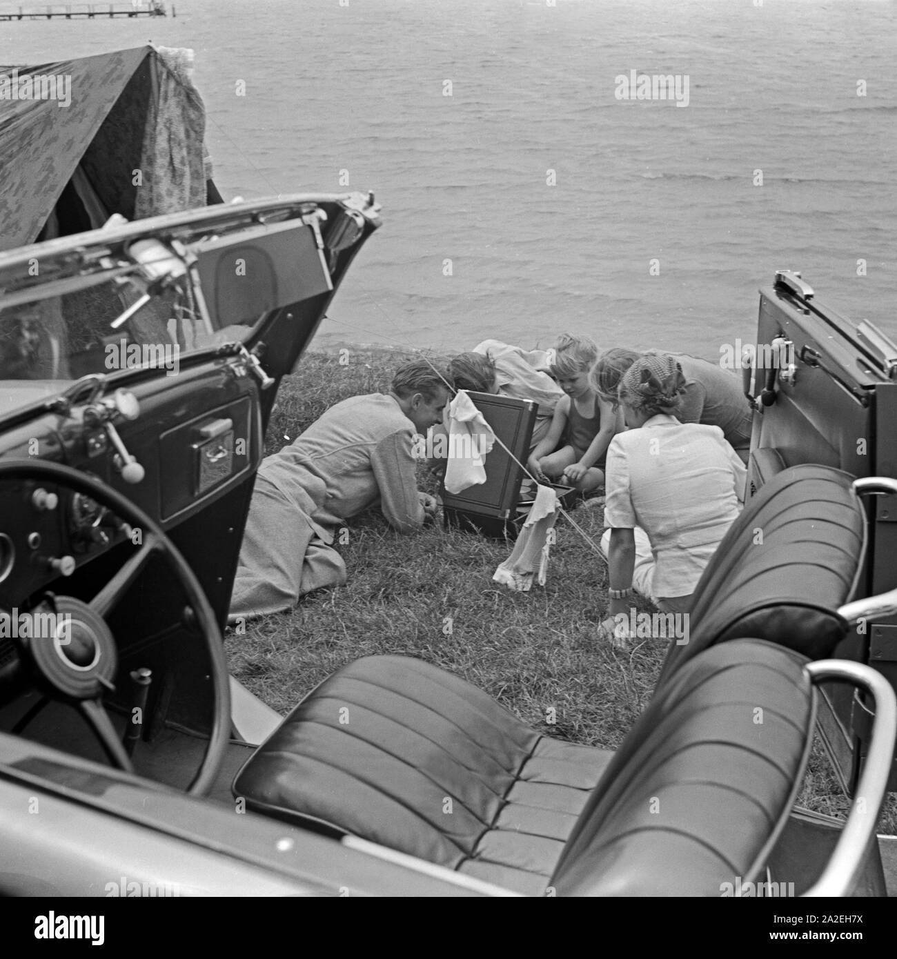 Campingausflug mit einem Ford V8 Cabrio ans Ufer eines Sees, wo die Familie um ein Electrole Koffer 106 Grammophon sitzt, Deutschland 1930er Jahre. Camping trip with a Ford V8 convertible to the shore of a lake, where the family is listening to music, played by an Electrola Koffer 106 suitcase gramophone, Germany 1930s. Stock Photo