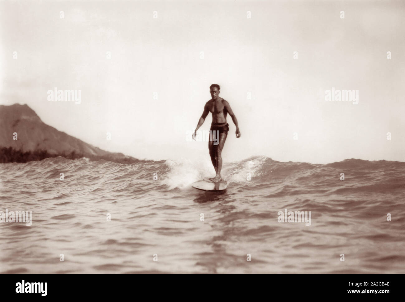 Olympic Gold Medal swimmer and Father of Modern Surfing, Duke Kahanamoku, riding a wave on a wooden surfboard in Waikiki, Hawaii with Diamond Head in the background. Stock Photo