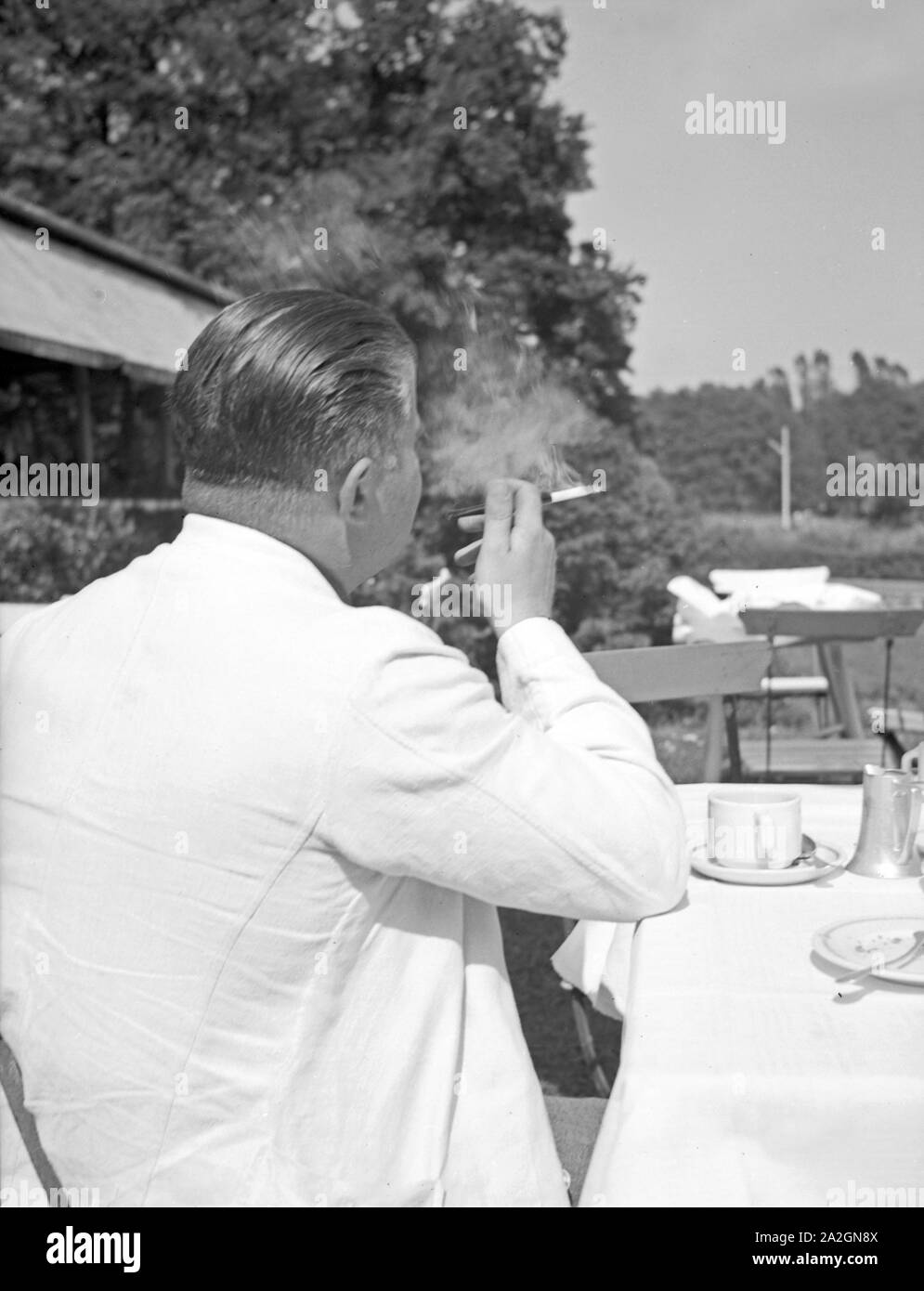 Ein Mann sitzt in einer Außengastronomie und raucht genüßlich eine Zigarette, Deutschland 1930er Jahre. A man sitting in the open air gastronomy of a restaurant smoking a cigarette, Germany 1930s. Stock Photo