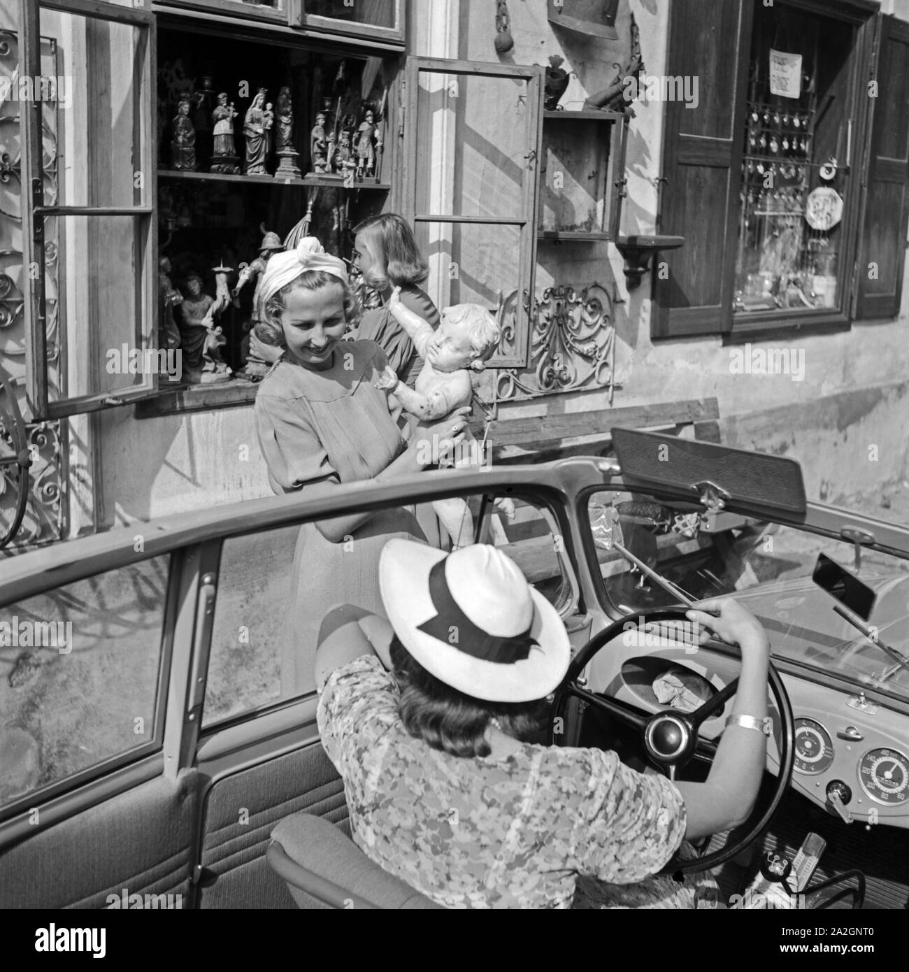 Zwei Frauen unterhalten sich aus dem Auto heraus vor einem Laden mit Kunsthandwerk, Deutschland 1930er Jahre. Two women chatting out from a convertible in front of a shop with craftwork, Germany 1930s. Stock Photo