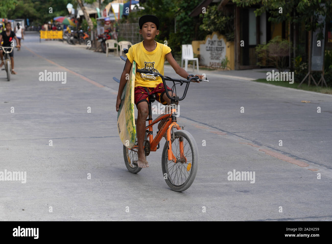 A young boy carrying a surfboard whilst riding a pedal cycle,Siargao,Philippines Stock Photo