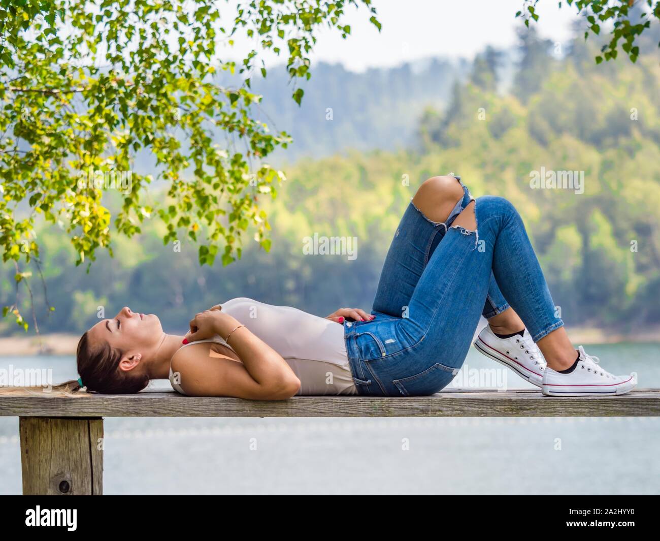 Young woman wearing Blue denim pants denims is resting lying on her back on wooden bench in park near lake country-side countryside in on at near but Stock Photo