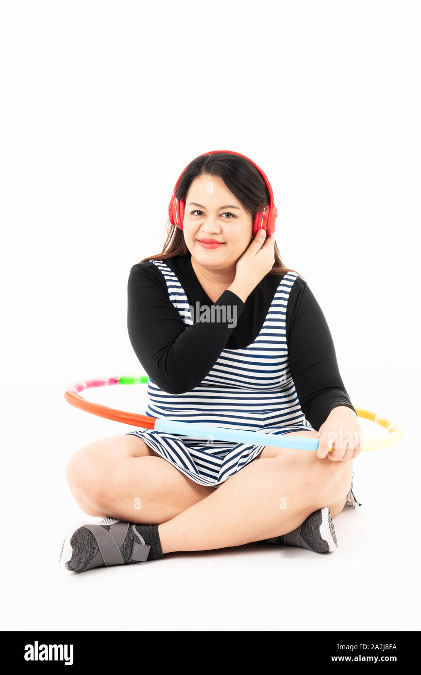 Fat woman resting and listening to music from red headphones after exercising by colorful Hula-Hoop. She smiles and enjoys losing weight for her bette Stock Photo