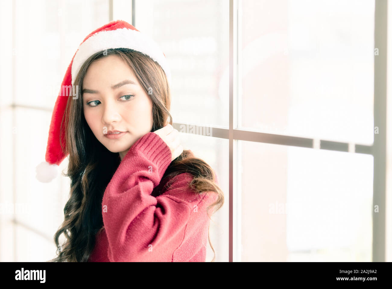 Close up one beautiful young Asian woman wearing a Santa hat is standing near the window on Christmas Day, waiting for a surprise gift from her boyfri Stock Photo