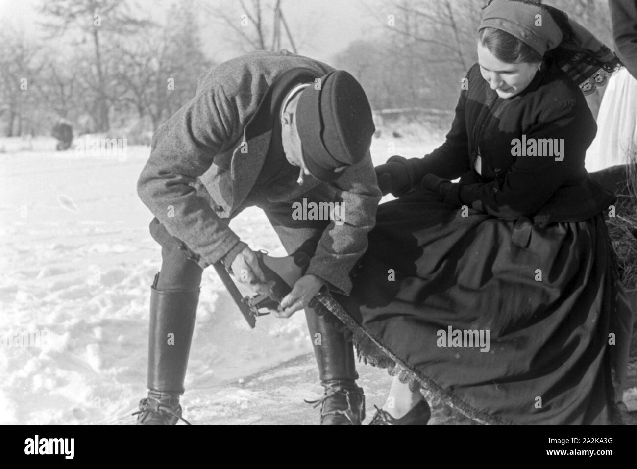 Ein Mann hilft einer Frau beim Anschnallen von Schlittschuhkufen im Spreewald, Deutschland 1930er Jahre. A man helping a woman to put on ice skate runners at Spreewald area, Germany 1930s. Stock Photo