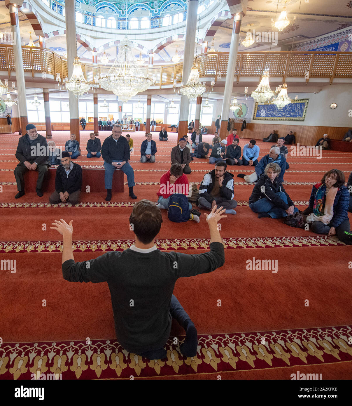Mannheim, Germany. 03rd Oct, 2019. Mikail Kibar, Deputy Chairman of the Turkish-Islamic Community of Mannheim e.V. (front), explains the prayer room of the Yavuz Sultan Selim Mosque to visitors on Open Mosque Day. Muslims are sitting in the background waiting for the midday prayer. Around 1300 mosques in Germany open their doors to interested guests. Credit: Tom Weller/dpa/Alamy Live News Stock Photo