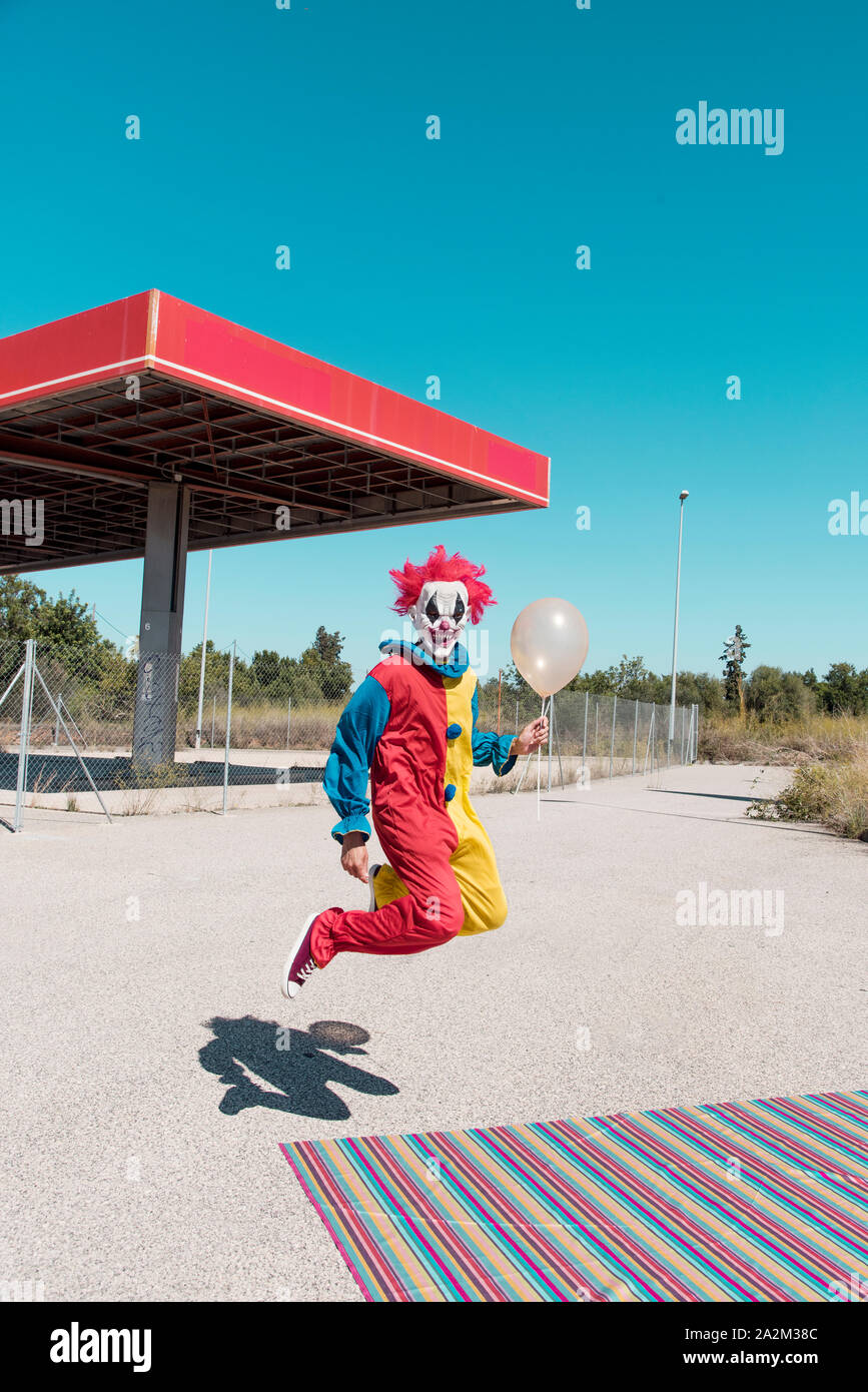 a scary clown wearing a colorful yellow, red and blue costume, holding a golden balloon in his hand, jumping in front of an abandoned filling station Stock Photo