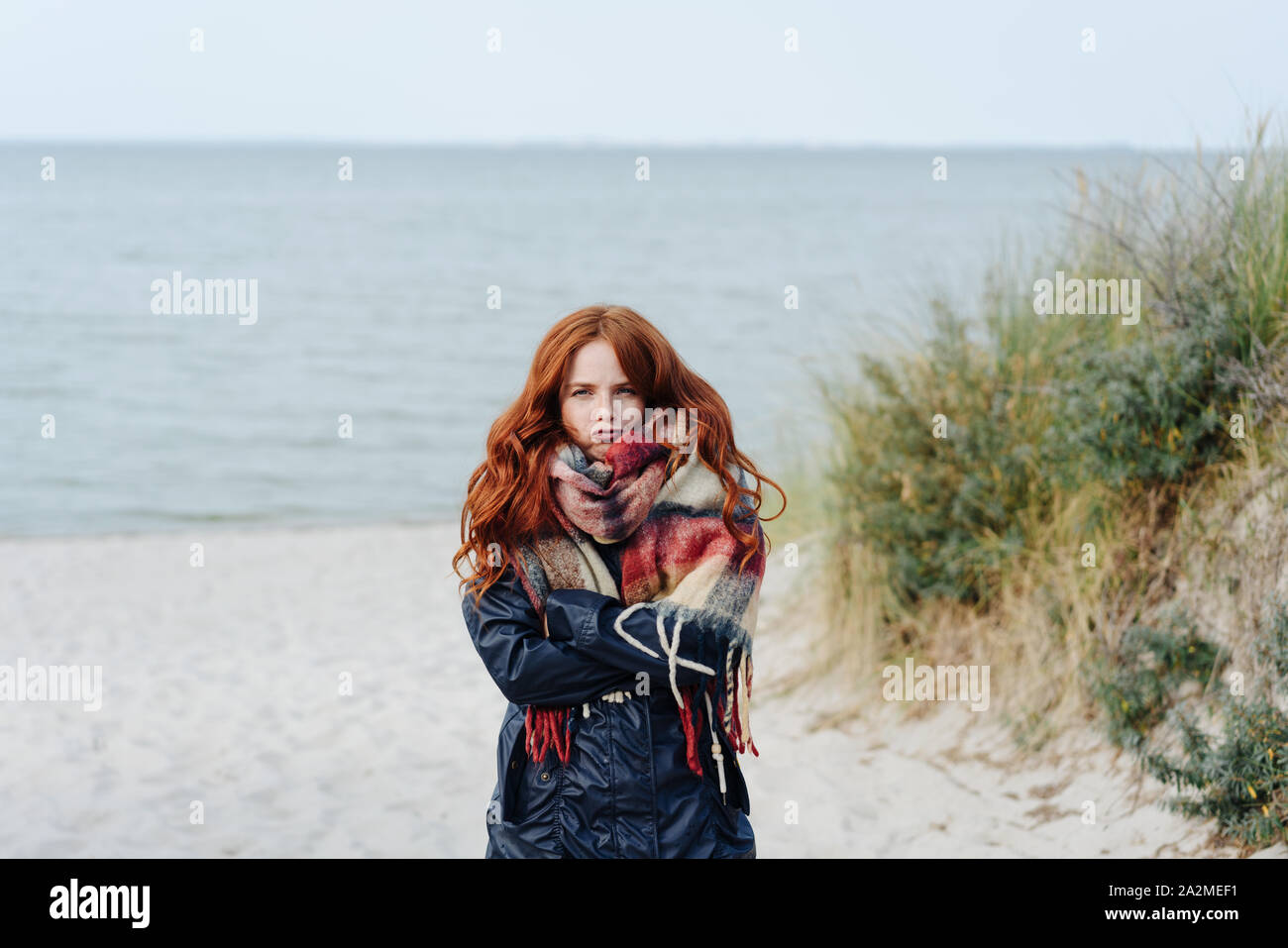 Unimpressed young woman shivering in the cold on a late autumn or winter day as she takes a walk on a sandy beach in thick anorak and scarf Stock Photo