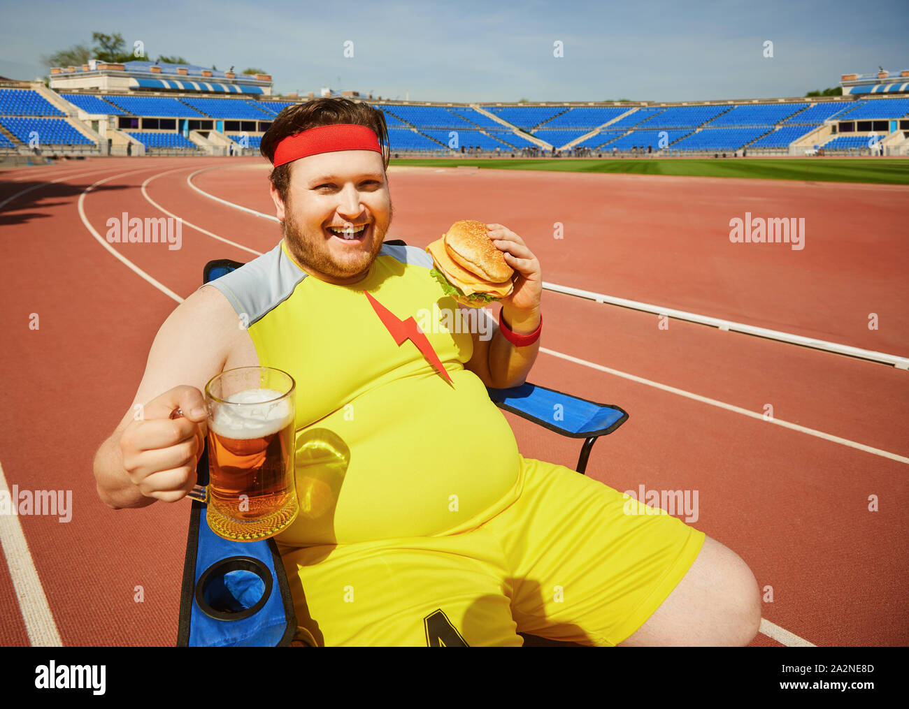 Fat man eating a burger and beer in training at the stadium. Stock Photo