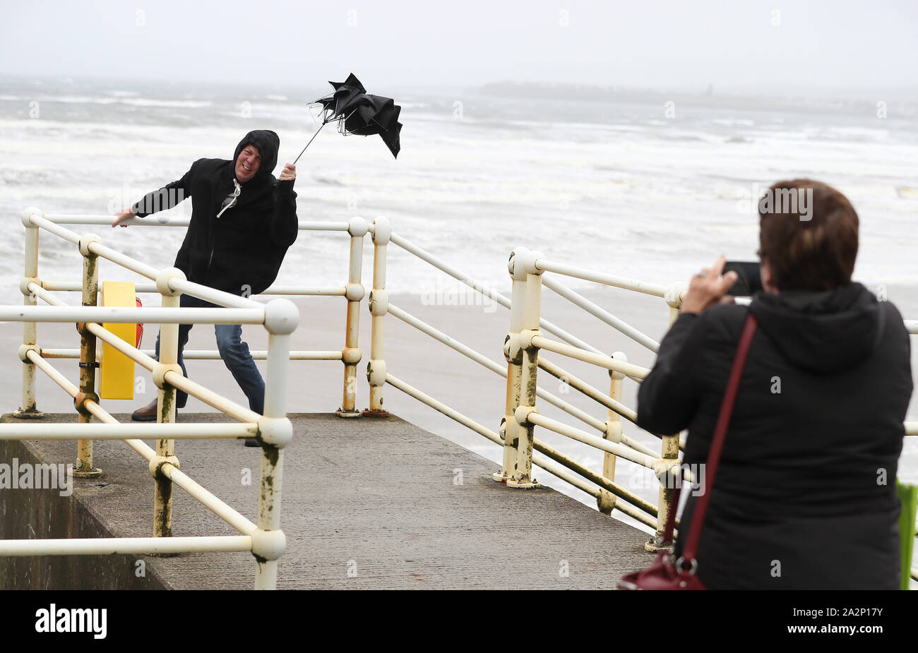 American tourists take photos with a broken umbrella along the sea front in Lahinch, County Clare, on the West Coast of Ireland as storm Lorenzo makes landfall, with a status orange wind warning and a yellow rain warning having been issued. Stock Photo