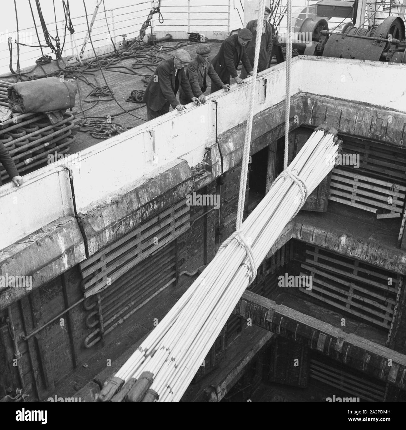 1950s, historical, London Docks, male dockworkers wearing flat caps overseeing heavy steel piping for export being lowered into the hull of a ship moored at the docks. Stock Photo