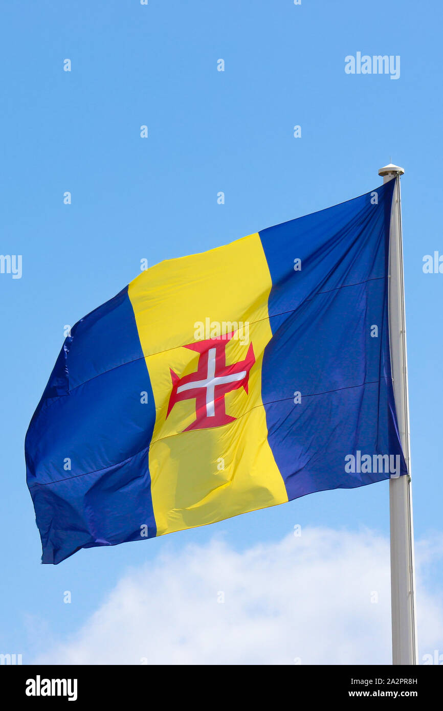 Waving flag of Madeira with blue sky in the background. It consists of a blue-yellow-blue vertical triband with a red-bordered white Cross of Christ in the center. Portuguese island in Atlantic ocean. Stock Photo